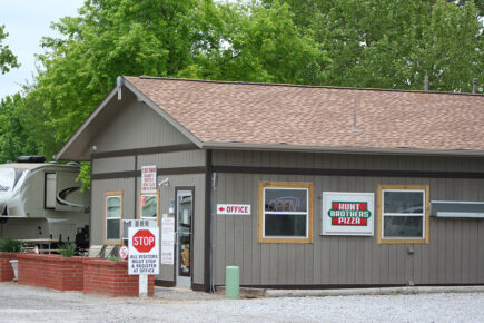 Small Building with Signs for "Office" and "Hunt Brothers Pizza," Surrounded by Trees, Gravel Lot, and an RV in the Background at Several RVs are Parked in a Wooded Campground Area with Trees and Picnic Tables Under a Clear Blue Sky at Council Road RV Park