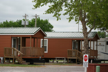 Two Small Brown Cabins with Porches that are Next to a Tree, Near an Office Sign, at Council Road RV Park