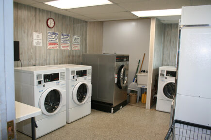 Laundry Room with Multiple Washers and Dryers, Cleaning Supplies, and a Wall Clock Above Signs on a Light-Colored Wall at Several RVs are Parked in a Wooded Campground Area with Trees and Picnic Tables Under a Clear Blue Sky at Council Road RV Park