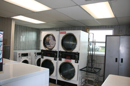 Interior of a Laundromat with Stacked Industrial Dryers and Washers, a Laundry Cart, and Fluorescent Lighting at Several RVs are Parked in a Wooded Campground Area with Trees and Picnic Tables Under a Clear Blue Sky at Council Road RV Park